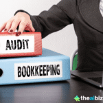 A person's hand reaching for a red binder labeled "AUDIT," with a blue binder labeled "BOOKKEEPING" underneath it, next to a laptop, symbolizing the organization of financial records enhanced by AI automated bookkeeping for small online shops.